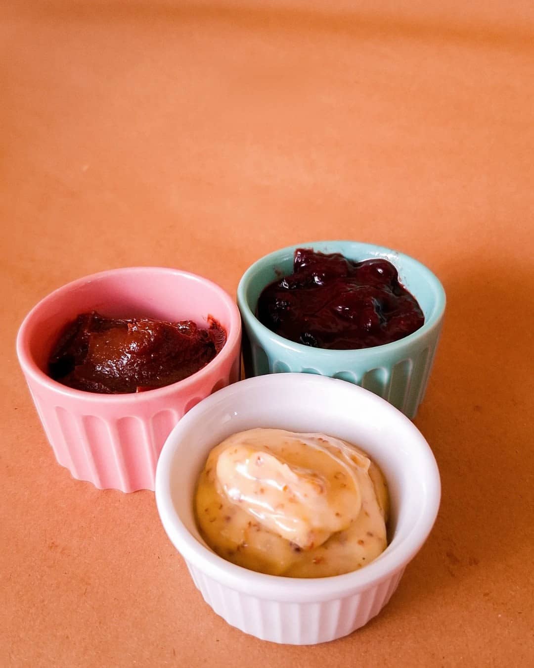 Três potinhos de cerâmica — rosa, verde-água e branco — com acompanhamentos doces e salgados para o croissant, sobre papel kraft.
