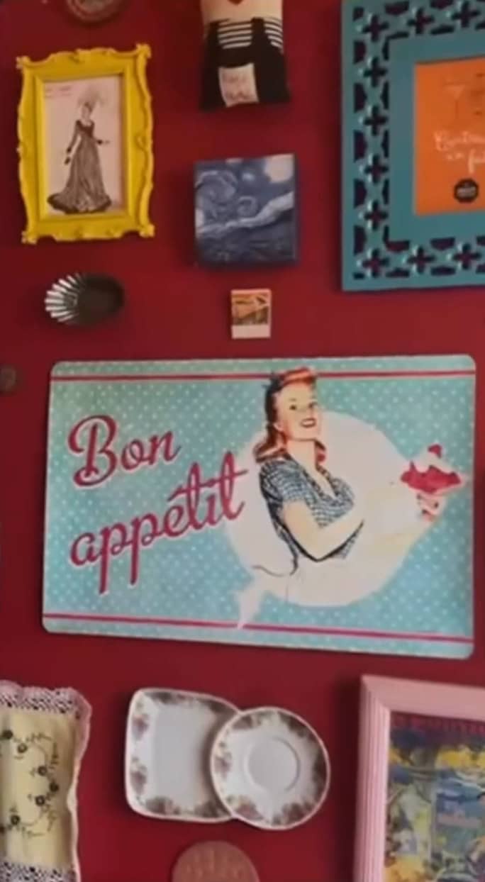 Placa “Bon appétit” com uma moça de avental, cercada de molduras, pratinhos e toalhinhas na parede vermelha.