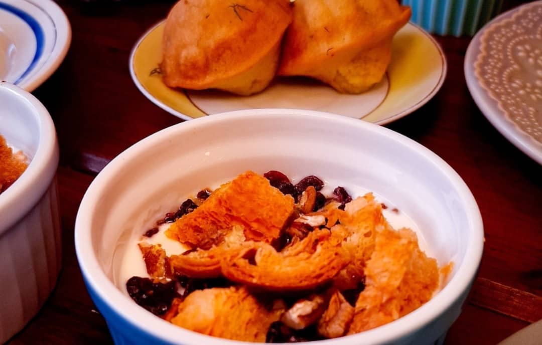 Potinho azul com creme, frutas secas e pedaços crocantes de croissant; madeleines ao fundo.